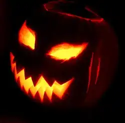 A jack-o'-lantern. The eyes and mouth of a carved pumpkina are lit from within while the rest of the carved vegetable is in darkness against a black background.