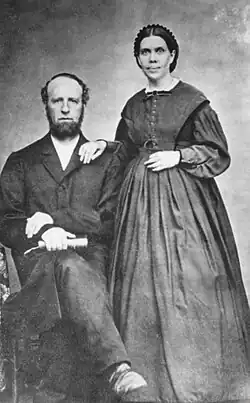 A balck and white photograph of a sitting balding man with a beard and a standing woman