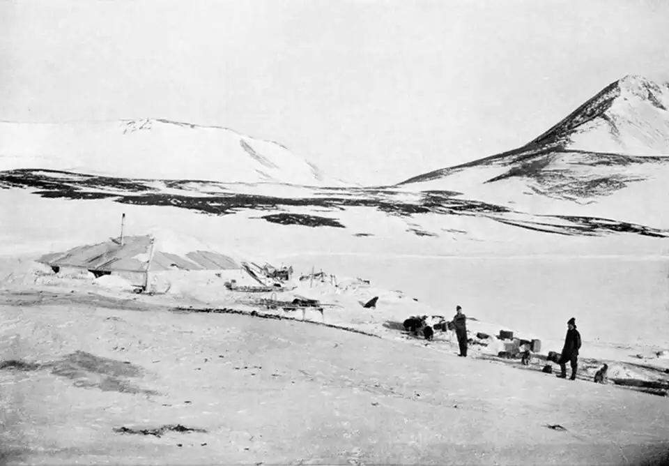 Photograph of two men in the middle distance standing amid supplies outside a low, snow-covered hut