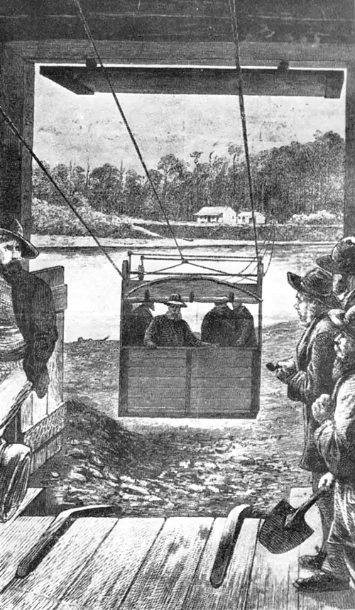 The “bird-cage” used to ferry people and supplies across the Taramakau River. It was a box slung on wire, with pulleys running on an overhead wire rope, and propelled by means of steam winches. The picture shows it on one side of the river with several men in the box, and men in the foreground at a terminal waiting their turn. On the other side of the river are two buildings with tall trees in the background.