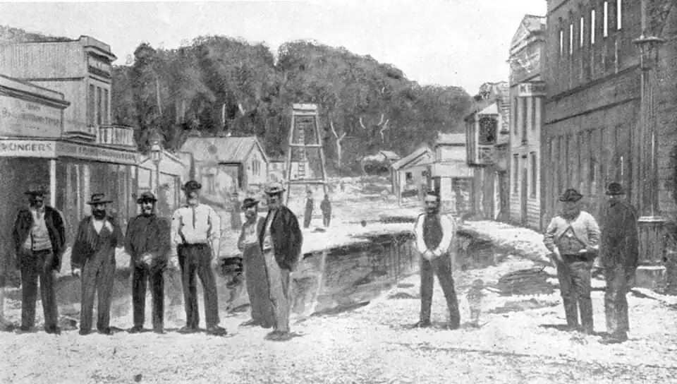 Wooden buildings on either side of a street (some two and three stories high) with bush and trees at the far end, and several men lined up across the street.