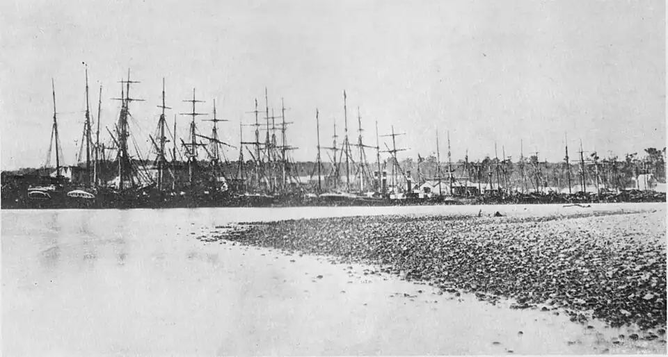 Several tall masted ships at port with the river in front and a gravel bank in the bottom right