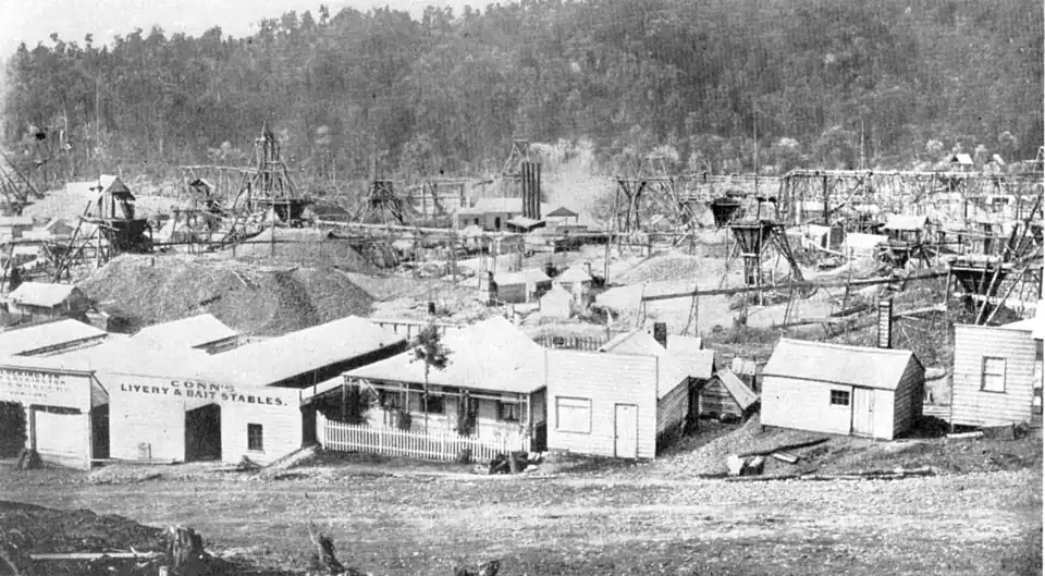 A flat plain with trees in the background going up a slope; in the foreground is a row of buildings, in the middle is an active gold field with piles of rock and equipment for extracting gold