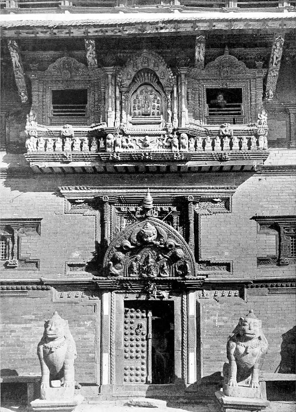 Black and white photograph of a door, surmounted by an ornamental lintel and flanked by statues on either side. The wall features two windows high above with an ornate balcony.