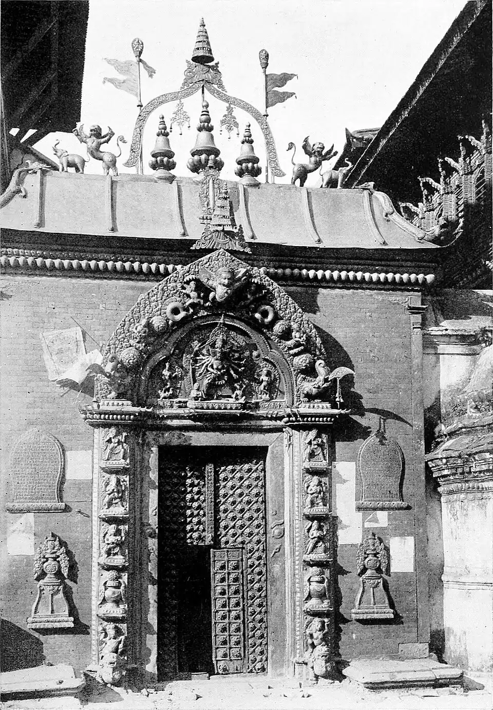 Black and white photograph of a large gate, which includes a smaller, partially open entrance way in the centre. The gate features a surface decoration of regular hemispheres. It is set between ornately carved pillars with an even more ornate lintel above. The sloped roof and the building can be seen at the top of the photograph.