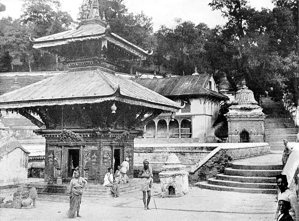 A black and white photograph of a village. a small three storey building, with two sloped roofs stands at different levels, to the left of the image. To the right, steps lead up to a higher area. A larger building can be seen in the background, at this higher level, in front of trees. People stand in the foreground.