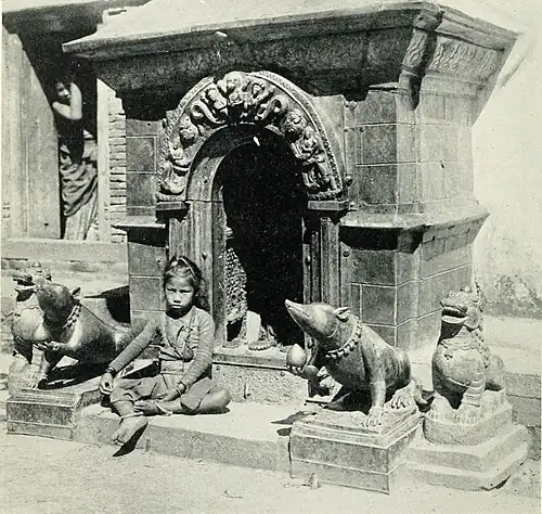 Black and white photograph of shrine dedicated to Ganesh at Patan. A small square stone structure with an arched entrance. Two animal statues are either side of the entrance, and a child sits on the step in front of it.