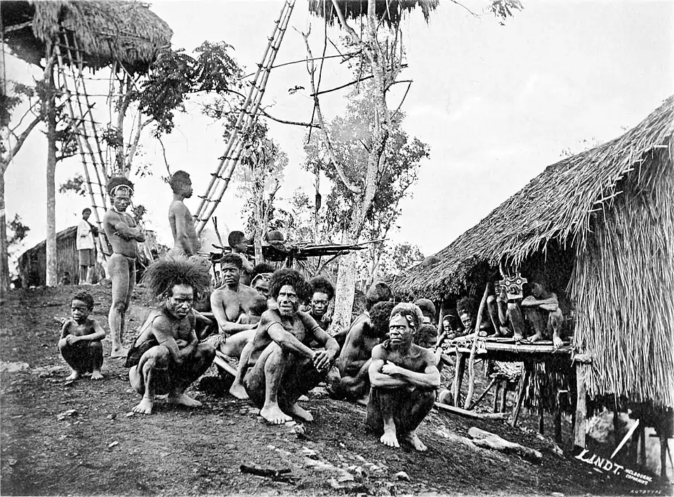 Black and white photograph of men crouching on the ground, next to a hut and trees.