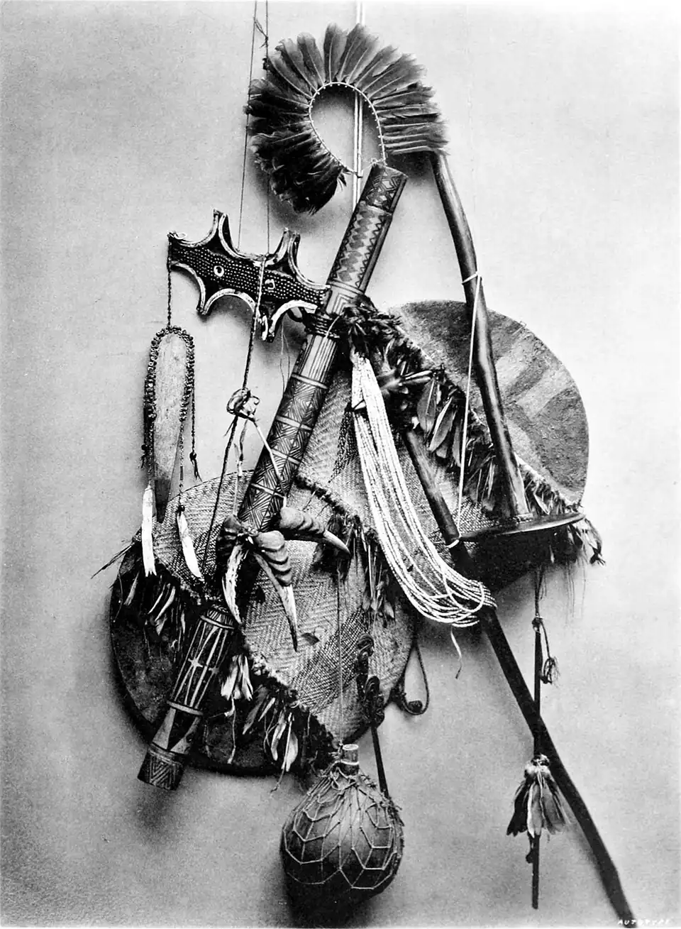 Black and white photograph of a collection of several implements. Includes, feathered headdress, an axe-like weapon and circular objects.