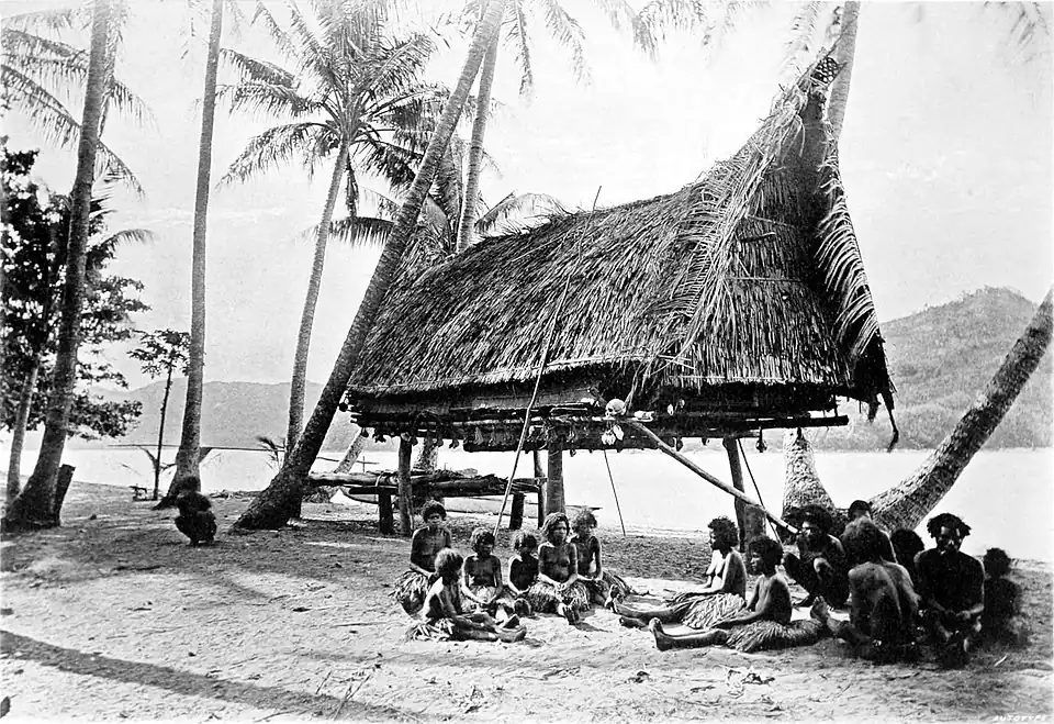 Black a white photograph of a thatched hut raised to head height among scattered trees on a beach. Two small groups of people sit in the foreground. Land can be seen on in the background, across the water.