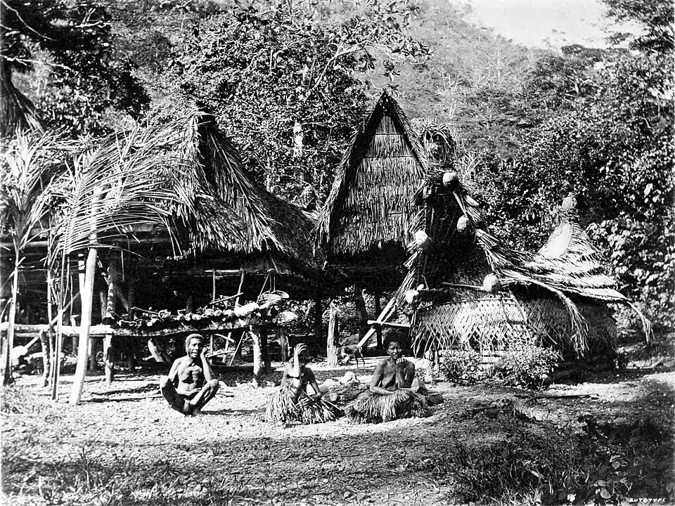Black and white photograph of a village of huts in a wooded area. Three people sit in the foreground.