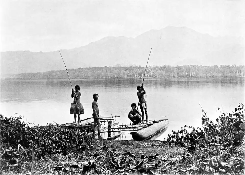 Black and white photograph of three men and a woman with a moored canoe. Land can be seen in the background, across the water, with a forest and distant mountains.