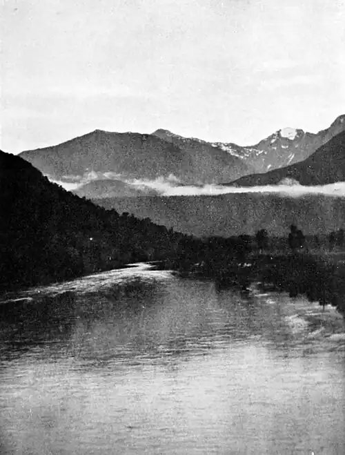 Calm river with bush a covered hill to the left and mist, hills and mountains with scattered snow in the distance