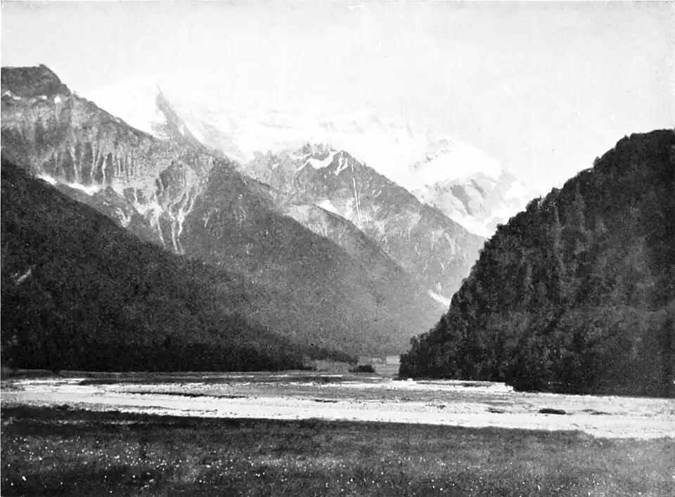 Flat plain before a river with steep hills on either side and snow-covered mountains in the background