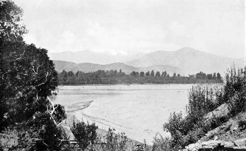Section of a river, with a bush covered bank on the far side and hills and mountains in the background.