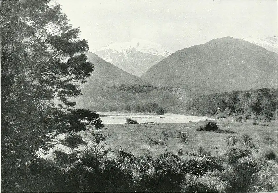 An open area before a river, with a tree ono the left-hand front side, the hills beyond the river meet in the center in a "V", with a snow-capped mountain in the center further in the distance