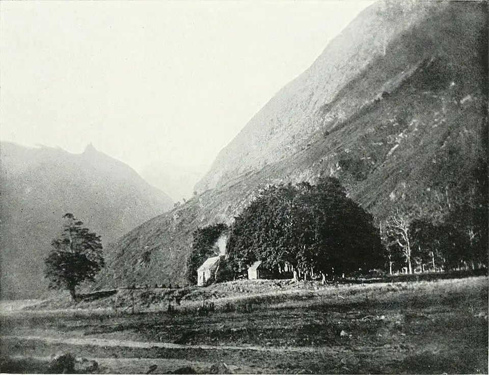 A very steep hill with a flat area n the front, at the base of the hill there is a clump of trees with a small house to the left with smoke coming out the chmney