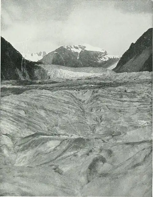 Undulating ice formation on the Fox Glacier, with steep hills on either side and snow-capped mountains in the distance