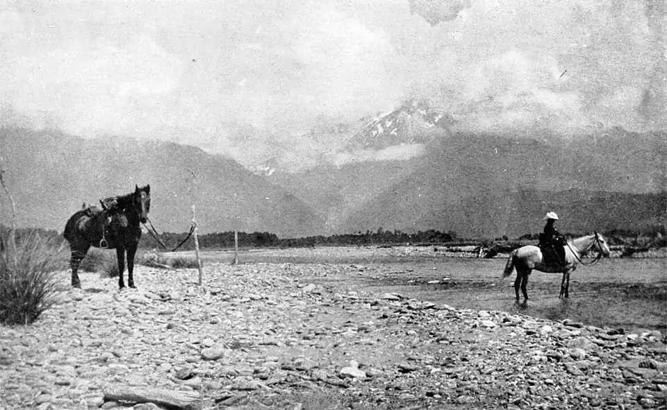 Rider on a horse crossing a river, with another horse hitched near the water's edge. The river has a stony bank and cloud covered mountains and hills in the background.