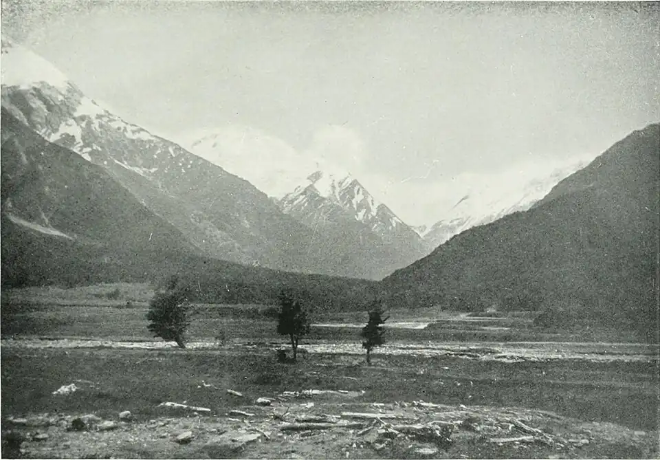 A plain with three trees spaced apart, with hills on either side and mountains in the background
