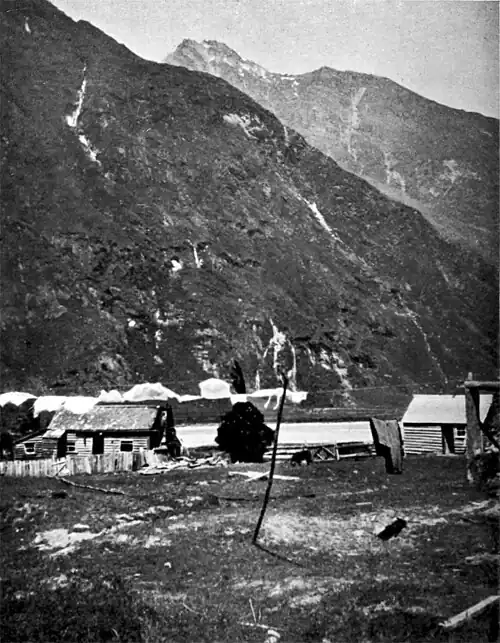 Two very steep mountains, one with a scattering of snow, with the river near the base and then on a flat grassy plain in front is a small house with a fence, a shed and tree.