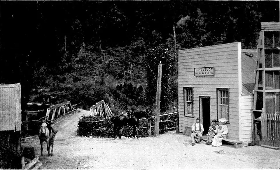 A road going towards a bridge surrounded by bush. A build is on the right with people sitting in front and tethered horses.