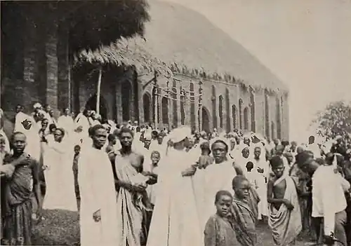 A large crowd gathered outside a church with a thatched roof, many of them in white robes