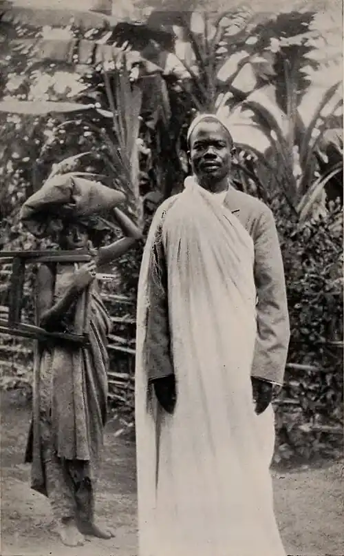 A man posing for the photo with a woman behind him carrying furniture and blankets