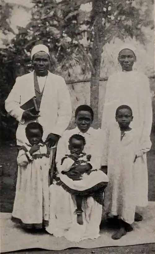 A man standing and holding a book, a woman standing to his side, and their several children seated in front of them, all wearing white