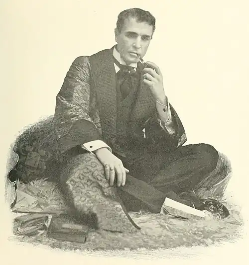 William Gillette as Sherlock Holmes, seated, wearing a dressing gown, and smoking a pipe.